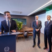 Discurso em ambiente institucional, com o governador posicionado atr&aacute;s de um p&uacute;lpito com emblema oficial e elemento gr&aacute;fico com cores da It&aacute;lia. Ao lado, outras pessoas permanecem em p&eacute; acompanhando a fala. 