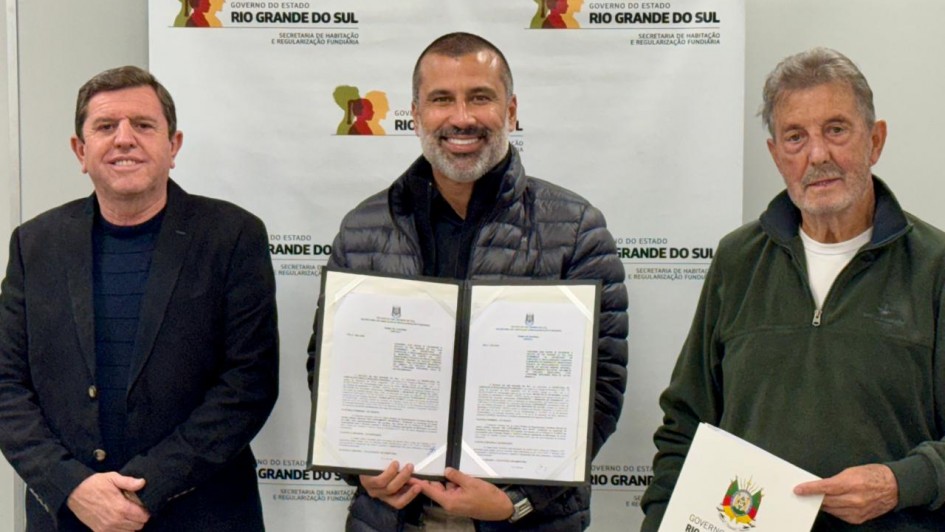 A imagem mostra um registro institucional em ambiente interno, com tr&ecirc;s pessoas posicionadas diante de um painel com a identidade do Governo do Estado do Rio Grande do Sul &ndash; Secretaria de Habita&ccedil;&atilde;o e Regulariza&ccedil;&atilde;o Fundi&aacute;ria. No centro, uma pessoa segura um porta-documentos aberto, exibindo dois termos oficiais impressos. 