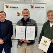 A imagem mostra um registro institucional em ambiente interno, com tr&ecirc;s pessoas posicionadas diante de um painel com a identidade do Governo do Estado do Rio Grande do Sul &ndash; Secretaria de Habita&ccedil;&atilde;o e Regulariza&ccedil;&atilde;o Fundi&aacute;ria. No centro, uma pessoa segura um porta-documentos aberto, exibindo dois termos oficiais impressos. 