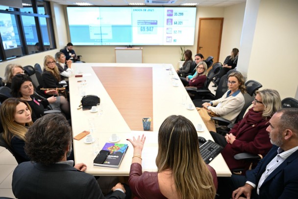 Em uma reunião com cerca de quinze pessoas em torno de uma mesa retangular. Um dos participantes, posicionado na ponta da mesa, parece conduzir a discussão.