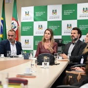 Em uma reuni&atilde;o oficial, v&aacute;rios participantes est&atilde;o sentados em torno de uma mesa de confer&ecirc;ncia, com copos, cadernos e objetos pessoais sobre a superf&iacute;cie. Ao fundo, um painel institucional exibe o nome da Secretaria da Seguran&ccedil;a P&uacute;blica e o bras&atilde;o do Estado, acompanhado das bandeiras do Brasil e do Rio Grande do Sul. A cena reflete um encontro administrativo voltado para pol&iacute;ticas de seguran&ccedil;a p&uacute;blica.