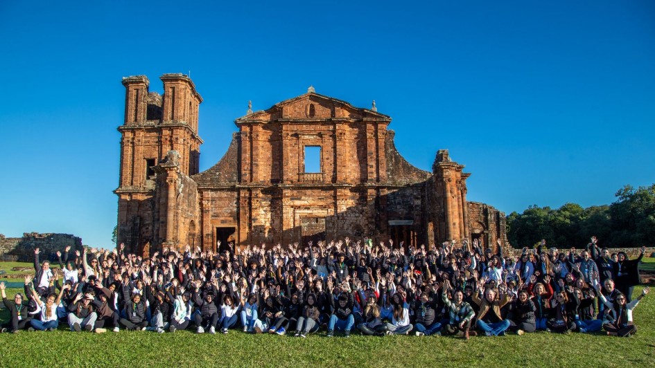 Registro coletivo ao ar livre em frente &agrave;s ru&iacute;nas hist&oacute;ricas das Miss&otilde;es, com grande grupo posicionado sobre gramado. A constru&ccedil;&atilde;o de pedra ocupa o fundo da cena, sob c&eacute;u azul limpo.