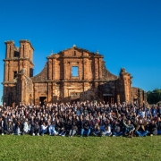 Registro coletivo ao ar livre em frente &agrave;s ru&iacute;nas hist&oacute;ricas das Miss&otilde;es, com grande grupo posicionado sobre gramado. A constru&ccedil;&atilde;o de pedra ocupa o fundo da cena, sob c&eacute;u azul limpo.