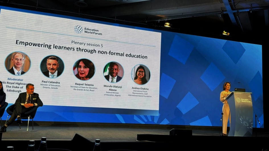 Painel em palco do Education World Forum, com tel&atilde;o exibindo a sess&atilde;o &ldquo;Plenary session 5 &ndash; Empowering learners through non-formal education&rdquo;. O slide apresenta fotos e nomes de participantes internacionais. &Agrave; direita do palco, a secret&aacute;ria da Educa&ccedil;&atilde;o est&aacute; em p&uacute;lpito com microfones e laptop, enquanto outro participante est&aacute; sentado &agrave; esquerda. 