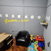 Sala de atendimento identificada como &ldquo;Sala das Margaridas&rdquo;, com decora&ccedil;&atilde;o acolhedora e tem&aacute;tica infantil. O espa&ccedil;o inclui poltrona, mesa de trabalho, brinquedos, tapete colorido e prateleira com objetos. Na parede, h&aacute; o texto &ldquo;Respeito e Prote&ccedil;&atilde;o&rdquo; em amarelo, al&eacute;m de flores decorativas.