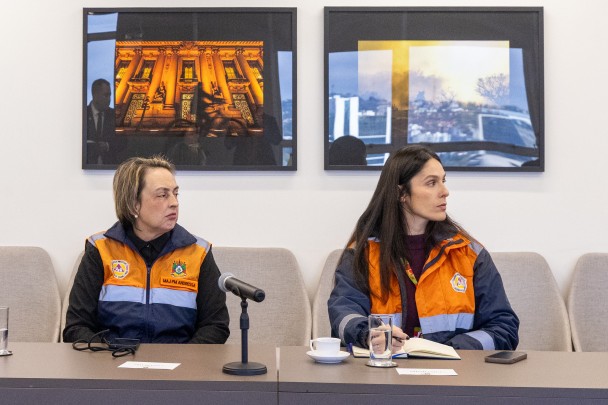 Foto mostra duas integrantes da Defesa Civil com coletes laranjas e azuis.