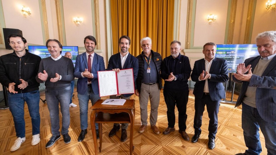Esta &eacute; uma descri&ccedil;&atilde;o da imagem "Governo do Estado assina ordem de in&iacute;cio para obras de recupera&ccedil;&atilde;o da ERS-437 e da RSC-453, na Serra.jpeg" para fins de acessibilidade:

A fotografia em plano inteiro mostra um grupo de oito homens alinhados lado a lado em um sal&atilde;o nobre de arquitetura cl&aacute;ssica, celebrando um ato oficial.

A&ccedil;&atilde;o Central: No centro do grupo, o governador Eduardo Leite segura e exibe uma pasta de capa vermelha aberta, apresentando o documento oficial assinado. &Agrave; sua frente, h&aacute; uma pequena mesa de madeira trabalhada (estilo bomb&ecirc;) com uma caneta sobre ela. Os demais homens ao redor sorriem e aplaudem o momento.

Os Participantes:

O governador Eduardo Leite (quarto da esquerda para a direita) veste um palet&oacute; azul-marinho, camisa branca sem gravata e cal&ccedil;a clara.

&Agrave; sua direita (esquerda da foto), encontram-se tr&ecirc;s homens: um de palet&oacute; azul e gravata bord&ocirc;, um de su&eacute;ter cinza-escuro e camisa clara, e um jovem de jaqueta preta e cal&ccedil;a jeans na extremidade.

&Agrave; sua esquerda (direita da foto), est&atilde;o quatro homens: um de colete acolchoado preto com um crach&aacute; oficial de cord&atilde;o colorido, um de casaco escuro, um de palet&oacute; azul-marinho e camisa clara, e um senhor de cabelos grisalhos e palet&oacute; xadrez azul na extremidade direita.

O Ambiente:

O sal&atilde;o possui um piso de parquet de madeira com desenhos geom&eacute;tricos bem marcados e brilhantes.

As paredes t&ecirc;m detalhes em tons claros e dourados, com arandelas de ilumina&ccedil;&atilde;o cl&aacute;ssicas fixadas.

Ao fundo, grandes cortinas de tecido pesado em tom dourado cobrem uma abertura.

Duas telas de TV dispostas nas laterais ao fundo exibem uma apresenta&ccedil;&atilde;o azul com o t&iacute;tulo do evento, onde se l&ecirc; parcialmente "ASSINATURA DE ORDEM DE IN&Iacute;CIO" e detalhes das rodovias como "ERS-437".

A imagem registra um momento formal de assinatura de investimentos em infraestrutura rodovi&aacute;ria com a presen&ccedil;a de lideran&ccedil;as pol&iacute;ticas.