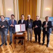 Esta &eacute; uma descri&ccedil;&atilde;o da imagem "Governo do Estado assina ordem de in&iacute;cio para obras de recupera&ccedil;&atilde;o da ERS-437 e da RSC-453, na Serra.jpeg" para fins de acessibilidade:

A fotografia em plano inteiro mostra um grupo de oito homens alinhados lado a lado em um sal&atilde;o nobre de arquitetura cl&aacute;ssica, celebrando um ato oficial.

A&ccedil;&atilde;o Central: No centro do grupo, o governador Eduardo Leite segura e exibe uma pasta de capa vermelha aberta, apresentando o documento oficial assinado. &Agrave; sua frente, h&aacute; uma pequena mesa de madeira trabalhada (estilo bomb&ecirc;) com uma caneta sobre ela. Os demais homens ao redor sorriem e aplaudem o momento.

Os Participantes:

O governador Eduardo Leite (quarto da esquerda para a direita) veste um palet&oacute; azul-marinho, camisa branca sem gravata e cal&ccedil;a clara.

&Agrave; sua direita (esquerda da foto), encontram-se tr&ecirc;s homens: um de palet&oacute; azul e gravata bord&ocirc;, um de su&eacute;ter cinza-escuro e camisa clara, e um jovem de jaqueta preta e cal&ccedil;a jeans na extremidade.

&Agrave; sua esquerda (direita da foto), est&atilde;o quatro homens: um de colete acolchoado preto com um crach&aacute; oficial de cord&atilde;o colorido, um de casaco escuro, um de palet&oacute; azul-marinho e camisa clara, e um senhor de cabelos grisalhos e palet&oacute; xadrez azul na extremidade direita.

O Ambiente:

O sal&atilde;o possui um piso de parquet de madeira com desenhos geom&eacute;tricos bem marcados e brilhantes.

As paredes t&ecirc;m detalhes em tons claros e dourados, com arandelas de ilumina&ccedil;&atilde;o cl&aacute;ssicas fixadas.

Ao fundo, grandes cortinas de tecido pesado em tom dourado cobrem uma abertura.

Duas telas de TV dispostas nas laterais ao fundo exibem uma apresenta&ccedil;&atilde;o azul com o t&iacute;tulo do evento, onde se l&ecirc; parcialmente "ASSINATURA DE ORDEM DE IN&Iacute;CIO" e detalhes das rodovias como "ERS-437".

A imagem registra um momento formal de assinatura de investimentos em infraestrutura rodovi&aacute;ria com a presen&ccedil;a de lideran&ccedil;as pol&iacute;ticas.