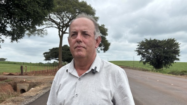 Foto mostra uma foto de perfil do produtor rural com a estrada ao fundo. 