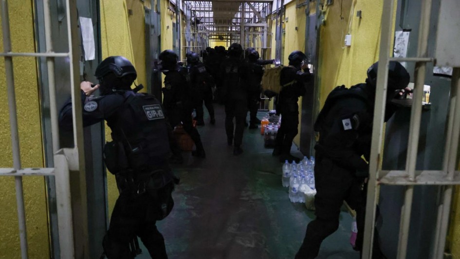 A imagem mostra o interior de um corredor institucional com portas met&aacute;licas laterais, onde diversos agentes uniformizados realizam uma a&ccedil;&atilde;o simult&acirc;nea, alguns abrindo portas e outros avan&ccedil;ando pelo espa&ccedil;o. 