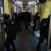 A imagem mostra o interior de um corredor institucional com portas met&aacute;licas laterais, onde diversos agentes uniformizados realizam uma a&ccedil;&atilde;o simult&acirc;nea, alguns abrindo portas e outros avan&ccedil;ando pelo espa&ccedil;o. 