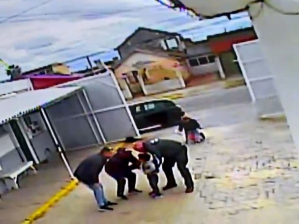 Foto mostra um momento captado por uma câmera de segurança. A cena mostra o pátio de uma instituição, com o portão aberto e um carro na entrada. O agente atende a criança que está com os pais. Outro servidor acompanha o atendimento.