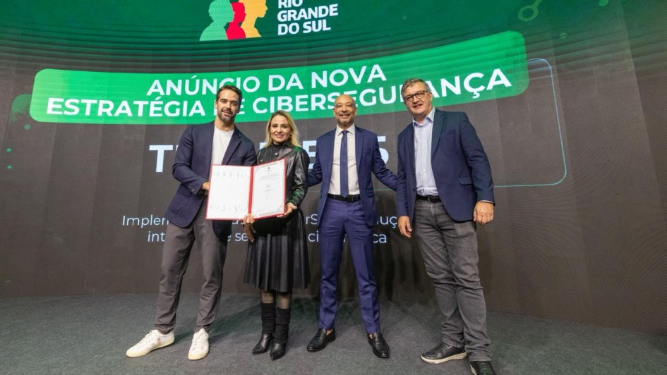 Esta &eacute; uma descri&ccedil;&atilde;o da imagem "Governo do Estado apresenta avan&ccedil;os do Programa RS+Digital e amplia parceria com munic&iacute;pios.jpeg" para fins de acessibilidade:

A fotografia em plano inteiro mostra quatro autoridades (tr&ecirc;s homens e uma mulher) alinhados lado a lado no centro de um palco, celebrando um an&uacute;ncio oficial.

A&ccedil;&atilde;o Central: &Agrave; esquerda, o governador Eduardo Leite e a mulher ao seu lado seguram juntos um documento oficial aberto em uma pasta de capa vermelha, exibindo-o para a c&acirc;mera. Todos os integrantes do grupo sorriem.

Os Participantes:

O governador Eduardo Leite (na extremidade esquerda) veste um palet&oacute; azul-marinho, camiseta branca, cal&ccedil;a cinza e t&ecirc;nis brancos.

A mulher ao seu lado veste um casaco longo de couro preto com saia plissada preta, meias escuras e botas pretas de cano m&eacute;dio.

O terceiro homem (centro-direita) veste um terno azul-vibrante com camisa branca, gravata escura e sapatos pretos sociais. Ele mant&eacute;m os bra&ccedil;os levemente abertos.

O quarto homem (na extremidade direita) usa &oacute;culos, veste um palet&oacute; azul-marinho sobre camisa azul-clara, cal&ccedil;a de sarja cinza e t&ecirc;nis escuros.

Plano de Fundo:

Atr&aacute;s do grupo, um grande tel&atilde;o de LED curvo exibe uma apresenta&ccedil;&atilde;o digital com fundo verde-escuro e detalhes gr&aacute;ficos.

No topo central do tel&atilde;o, destaca-se o logotipo oficial do Governo do Estado do Rio Grande do Sul (com o perfil de tr&ecirc;s rostos coloridos em verde, vermelho e amarelo).

Abaixo do logotipo, uma grande faixa verde-clara exibe em letras brancas o t&iacute;tulo: "AN&Uacute;NCIO DA NOVA ESTRAT&Eacute;GIA DE CIBERSEGURAN&Ccedil;A".

Mais abaixo,文字 brancas menores trazem textos informativos parcialmente vis&iacute;veis.

O Ambiente: O palco possui um revestimento cinza-escuro fosco e uniforme, com ilumina&ccedil;&atilde;o frontal direcionada para o grupo.

A imagem registra um momento institucional de avan&ccedil;o tecnol&oacute;gico e seguran&ccedil;a digital para o estado.