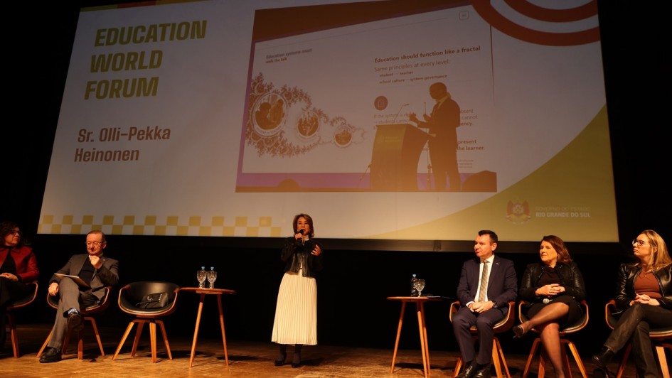 Esta &eacute; uma descri&ccedil;&atilde;o da imagem "Semin&aacute;rio do F&oacute;rum Permanente de Apoio &agrave; Forma&ccedil;&atilde;o de Professores fortalece pol&iacute;ticas de forma&ccedil;&atilde;o de professores no Estado.jpg" para fins de acessibilidade:

A fotografia em plano geral e &acirc;ngulo frontal mostra um painel de discuss&atilde;o em um palco de teatro ou audit&oacute;rio, com destaque para uma grande tela de proje&ccedil;&atilde;o ao fundo.

A Palestrante: No centro do palco, uma mulher de cabelos castanhos curtos est&aacute; de p&eacute; e fala ao microfone. Ela veste uma jaqueta de couro preta sobre blusa clara e uma saia longa plissada branca.

Os Integrantes do Painel:

&Agrave; direita, sentados em poltronas individuais de madeira com estofado escuro, tr&ecirc;s pessoas acompanham a apresenta&ccedil;&atilde;o: um homem de terno azul-escuro, camisa clara e gravata; uma mulher de casaco e saia escura com as pernas cruzadas; e uma mulher de jaqueta de couro preta e cal&ccedil;a escura.

&Agrave; esquerda, outras tr&ecirc;s pessoas tamb&eacute;m est&atilde;o acomodadas em poltronas semelhantes: um homem de palet&oacute; cinza que analisa pap&eacute;is em seu colo, uma mulher de blazer bord&ocirc; (parcialmente vis&iacute;vel na extremidade esquerda) e uma poltrona vazia com uma bolsa preta sobre o assento entre eles.

A Tela de Proje&ccedil;&atilde;o: O grande tel&atilde;o ao fundo exibe um slide institucional claro.

No lado esquerdo, l&ecirc;-se em letras mai&uacute;sculas douradas: "EDUCATION WORLD FORUM", seguido pelo nome "Sr. Olli-Pekka Heinonen" em marrom.

No centro, h&aacute; a reprodu&ccedil;&atilde;o de um slide em ingl&ecirc;s com o t&iacute;tulo "Education should function like a fractal", exibindo a silhueta de um homem discursando em um p&uacute;lpito e diagramas circulares interconectados.

No canto inferior direito do tel&atilde;o, consta o bras&atilde;o e o logotipo oficial do Governo do Estado do Rio Grande do Sul.

O Ambiente: O palco possui piso de t&aacute;buas de madeira claras e ilumina&ccedil;&atilde;o c&ecirc;nica direcionada. Pequenas mesas redondas de madeira servem de apoio para ta&ccedil;as e garrafas de &aacute;gua entre os participantes. O restante do cen&aacute;rio ao fundo &eacute; escuro.

A imagem registra um evento oficial e acad&ecirc;mico de relev&acirc;ncia internacional na &aacute;rea da educa&ccedil;&atilde;o.