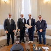 A imagem mostra quatro pessoas em p&eacute; em uma sala elegante, organizadas lado a lado em frente a uma grande janela com cortinas claras e luz natural entrando. Duas das pessoas ao centro seguram um documento, exibindo-o para a c&acirc;mera.