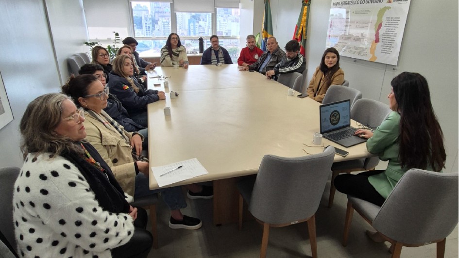 A imagem mostra uma reuni&atilde;o em uma sala de escrit&oacute;rio, com pessoas sentadas ao redor de uma mesa retangular grande e clara. Uma pessoa est&aacute; posicionada na cabeceira, utilizando um notebook aberto para apresentar informa&ccedil;&otilde;es ao grupo, enquanto os demais acompanham.