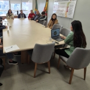 A imagem mostra uma reuni&atilde;o em uma sala de escrit&oacute;rio, com pessoas sentadas ao redor de uma mesa retangular grande e clara. Uma pessoa est&aacute; posicionada na cabeceira, utilizando um notebook aberto para apresentar informa&ccedil;&otilde;es ao grupo, enquanto os demais acompanham.