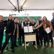 A fotografia em plano inteiro mostra um grupo numeroso de pessoas reunidas no centro de um palco sob uma estrutura de tenda branca, celebrando a assinatura de um ato oficial.

A&ccedil;&atilde;o Central: No centro do grupo, o governador Eduardo Leite segura uma pasta aberta de cor verde, exibindo um documento oficial com assinaturas. V&aacute;rias pessoas ao seu redor aproximam-se sorrindo para posar juntas para a foto. &Agrave; frente do governador, h&aacute; um pequeno balc&atilde;o ou p&uacute;lpito m&oacute;vel de cor clara.

Os Participantes:

O governador Eduardo Leite (ao centro) veste camisa branca sob um colete acolchoado preto com uma pequena bandeira do Rio Grande do Sul no peito, cal&ccedil;a escura e t&ecirc;nis claros.

&Agrave; sua direita (esquerda da foto), destacam-se uma mulher &agrave; frente usando um casaco preto com um cachecol estampado em tons de bege e preto, um homem na extremidade esquerda vestindo jaqueta acolchoada preta e cal&ccedil;a jeans, e uma mulher loira sorridente com colete acolchoado preto logo atr&aacute;s. Outras pessoas completam o grupo ao fundo.

&Agrave; sua esquerda (direita da foto), uma mulher com casaco marrom claro se inclina levemente segurando folhas de papel branco, ladeada por um homem de palet&oacute; cinza sobre camisa escura. Na extremidade direita, um homem robusto veste uma jaqueta acolchoada azul-marinho e cal&ccedil;a escura. Outros homens de trajes sociais e casuais acompanham o ato logo atr&aacute;s do p&uacute;lpito.

O Palco e Plano de Fundo:

O piso do palco &eacute; revestido por uma superf&iacute;cie verde que simula grama sint&eacute;tica.

Atr&aacute;s do grupo, h&aacute; um grande painel digital com fundo verde-brilhante.

Logo acima do painel, na lona branca da tenda, est&atilde;o aplicados os logotipos oficiais da CUFA (Central &Uacute;nica das Favelas), da Secretaria do Esporte e Lazer e do Governo do Estado do Rio Grande do Sul. No teto da estrutura, refletores de ilumina&ccedil;&atilde;o c&ecirc;nica aparecem fixados.

Na extrema esquerda, em segundo plano, nota-se uma bola de v&ocirc;lei ou futebol de cor amarela e azul sobre caixas de papel&atilde;o institucionais.

A imagem registra o momento de celebra&ccedil;&atilde;o e formaliza&ccedil;&atilde;o de parcerias entre o governo estadual, entidades sociais e representantes do esporte.