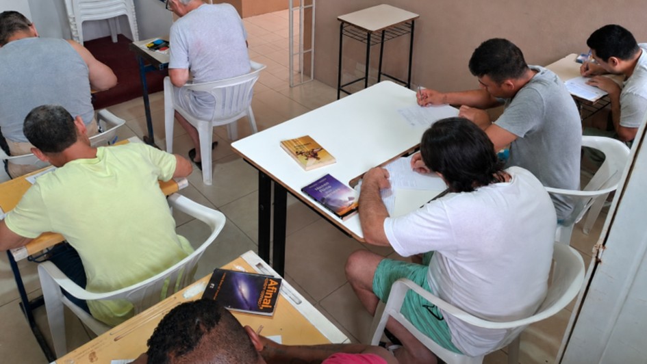 Foto mostra de cima uma sala onde h&aacute; apenados sentados em carteiras de sala de aula. Eles est&atilde;o lendo livros.
