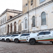 A imagem mostra diversas caminhonetes brancas adesivadas com a marca da Secretaria da Sa&uacute;de do RS, alinhadas em frente a um pr&eacute;dio hist&oacute;rico. 