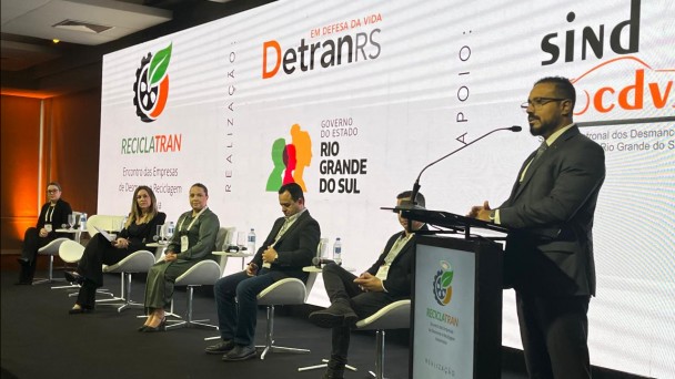 Foto mostra palestrante durante o evento, ele fala no p&uacute;lpito. Outros palestrantes est&atilde;o sentados acompanhando a fala. Ao fundo, um painel branco com o nome do evento e as logos do governo do Estado, do DetranRS e outros apoiadores do evento.