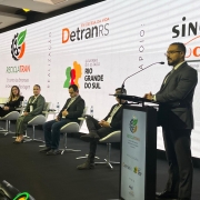 Foto mostra palestrante durante o evento, ele fala no p&uacute;lpito. Outros palestrantes est&atilde;o sentados acompanhando a fala. Ao fundo, um painel branco com o nome do evento e as logos do governo do Estado, do DetranRS e outros apoiadores do evento.