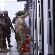 Corredor interno com paredes claras e ilumina&ccedil;&atilde;o homog&ecirc;nea, onde um grupo de agentes t&aacute;ticos usando uniformes camuflados, capacetes e equipamentos de prote&ccedil;&atilde;o conduz uma a&ccedil;&atilde;o em frente a uma fileira de portas met&aacute;licas de uma pris&atilde;o. 