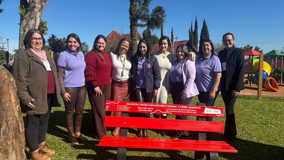A fotografia em plano inteiro mostra um grupo de nove mulheres posando sorridentes, alinhadas lado a lado em uma pra&ccedil;a p&uacute;blica ao ar livre durante um dia ensolarado. Elas est&atilde;o posicionadas logo atr&aacute;s de um banco de pra&ccedil;a pintado de vermelho vivo.

O Banco de Destaque:

Em primeiro plano, centralizado na metade inferior da foto, h&aacute; um banco de madeira com estrutura de apoio preta e assento e encosto pintados de vermelho.

No topo do encosto do banco, uma faixa adesiva vermelha exibe mensagens de conscientiza&ccedil;&atilde;o em letras brancas, onde se l&ecirc; em destaque: "VIOL&Ecirc;NCIA CONTRA A MULHER N&Atilde;O &Eacute; NORMAL. &Eacute; CRIME. DENUNCIE!". Textos menores trazem canais de den&uacute;ncia, telefones &uacute;teis e frases de apoio.

As Integrantes: As nove mulheres vestem roupas variadas, incluindo casacos de inverno, blusas de gola alta e camisas polo de uniformes institucionais.

Tr&ecirc;s delas usam camisas polo lil&aacute;s (duas &agrave; esquerda e uma mais &agrave; direita).

As demais vestem casacos em tons de marrom, vermelho-escuro, branco e preto.

Elas est&atilde;o abra&ccedil;adas ou posicionadas muito pr&oacute;ximas, transmitindo um sentimento de uni&atilde;o e parceria. Algumas usam crach&aacute;s oficiais pendurados no pesco&ccedil;o.

O Ambiente:

O grupo est&aacute; sobre um gramado verde bem cuidado e iluminado pelo sol direto.

No canto esquerdo, em primeir&iacute;ssimo plano, aparece a lateral de uma grande rocha ou tronco de &aacute;rvore texturizado.

Ao fundo, a pra&ccedil;a conta com &aacute;rvores altas (incluindo uma arauc&aacute;ria centralizada atr&aacute;s do grupo), ciprestes finos e a estrutura de um parquinho infantil colorido (com escorregadores em azul, amarelo e laranja).

O c&eacute;u ao fundo est&aacute; completamente limpo e azul, indicando um dia de clima firme.

A imagem registra uma a&ccedil;&atilde;o comunit&aacute;ria e institucional de conscientiza&ccedil;&atilde;o e engajamento no combate &agrave; viol&ecirc;ncia contra a mulher.