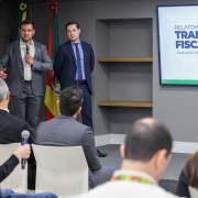 Sala de apresenta&ccedil;&atilde;o institucional com paredes neutras e ilumina&ccedil;&atilde;o uniforme, onde duas pessoas em trajes formais est&atilde;o em frente a um p&uacute;blico sentado. Ao lado, uma tela exibe o t&iacute;tulo &ldquo;RELAT&Oacute;RIO DE TRANSPAR&Ecirc;NCIA FISCAL // DATA BASE: 1Q2026 // EDI&Ccedil;&Atilde;O 20&rdquo;, acompanhado do logotipo do Governo do Estado do Rio Grande do Sul.