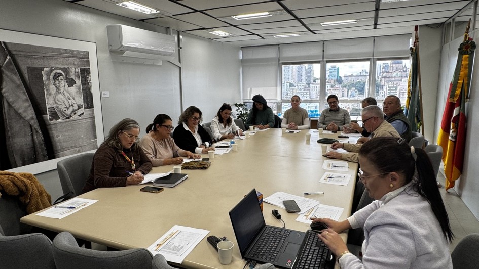 Esta &eacute; uma descri&ccedil;&atilde;o da imagem "Comiss&atilde;o Estadual define can&ccedil;&atilde;o-tema dos Festejos Farroupilhas 2026.jpg" para fins de acessibilidade:

A fotografia em plano geral mostra uma reuni&atilde;o de trabalho institucional acontecendo em uma sala de reuni&otilde;es corporativa de formato retangular.

A Mesa de Reuni&atilde;o: No centro da imagem, estende-se uma grande mesa retangular de cor bege clara, cercada por cadeiras estofadas de cor cinza-escura. Cerca de doze pessoas est&atilde;o acomodadas ao redor da mesa.

Os Participantes:

Em primeiro plano, no canto inferior direito, uma mulher de cabelos escuros presos em um rabo de cavalo e vestindo um casaco claro trabalha em um notebook preto aberto, utilizando um teclado num&eacute;rico auxiliar.

Ao longo do lado esquerdo da mesa, uma fileira de cinco mulheres est&aacute; sentada. A primeira delas, mais pr&oacute;xima &agrave; c&acirc;mera, usa &oacute;culos e casaco marrom enquanto faz anota&ccedil;&otilde;es em um papel. As demais aparecem concentradas, lendo documentos ou escrevendo.

Ao fundo e do lado direito da mesa, est&atilde;o sentados outros integrantes do grupo, incluindo homens e mulheres. Alguns conversam entre si e outros observam a reuni&atilde;o. Sobre a mesa, h&aacute; folhas com planilhas, canetas, smartphones e copos pl&aacute;sticos descart&aacute;veis.

O Ambiente:

A sala possui paredes de cor cinza-clara. Na parede da esquerda, h&aacute; um aparelho de ar-condicionado do tipo split desligado e um grande quadro decorativo com uma fotografia em preto e branco que retrata uma pessoa de perfil em um ambiente r&uacute;stico.

Ao fundo, grandes janelas de vidro com persianas integradas revelam uma vista panor&acirc;mica de uma &aacute;rea urbana com pr&eacute;dios e relevos sob um c&eacute;u nublado.

No canto superior direito, na cabeceira da mesa, est&atilde;o dispostas duas bandeiras oficiais em seus mastros, incluindo a bandeira do Estado do Rio Grande do Sul (com suas listras diagonais em verde, vermelho e amarelo).

O teto &eacute; composto por placas modulares com lumin&aacute;rias embutidas de luz branca, que iluminam todo o espa&ccedil;o de forma uniforme.

A imagem registra uma din&acirc;mica de planejamento e delibera&ccedil;&atilde;o coletiva em um ambiente administrativo governamental.