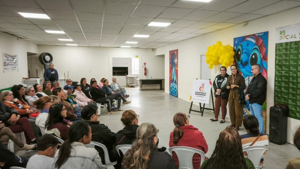 Foto mostra instrutores, em p&eacute;, falando em frente a plateia que est&aacute; sentada. A cena ocorre dentro de uma sala ampla.