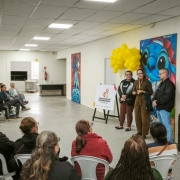Foto mostra instrutores, em p&eacute;, falando em frente a plateia que est&aacute; sentada. A cena ocorre dentro de uma sala ampla.