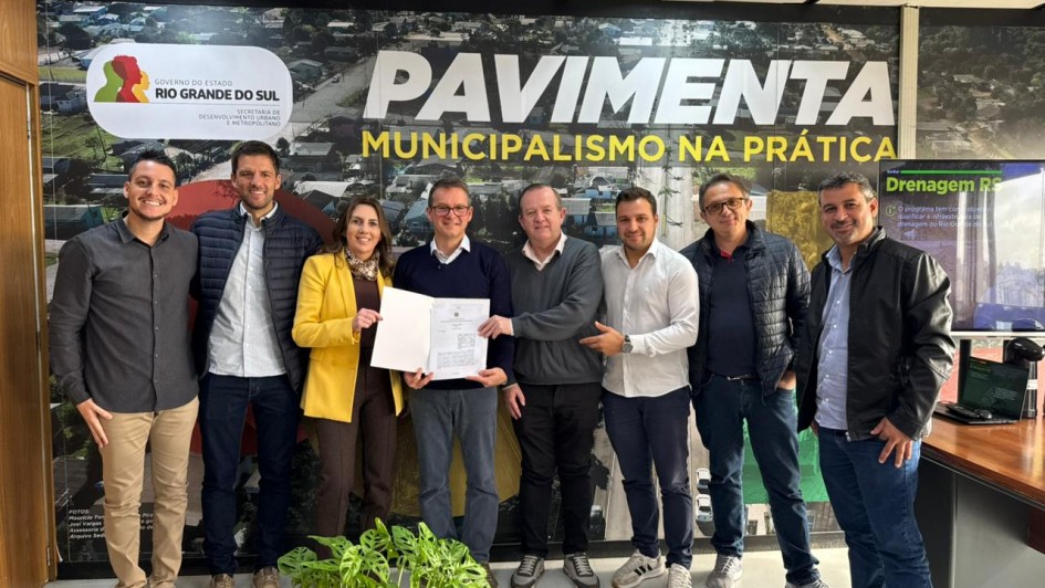 Ambiente interno de escrit&oacute;rio com ilumina&ccedil;&atilde;o de teto e mobili&aacute;rio simples, onde um grupo de sete pessoas posa lado a lado diante de um painel grande com a inscri&ccedil;&atilde;o &ldquo;PAVIMENTA &ndash; MUNICIPALISMO NA PR&Aacute;TICA&rdquo; e o logotipo do Governo do Estado do Rio Grande do Sul. No centro, duas pessoas seguram um documento aberto voltado para a c&acirc;mera.