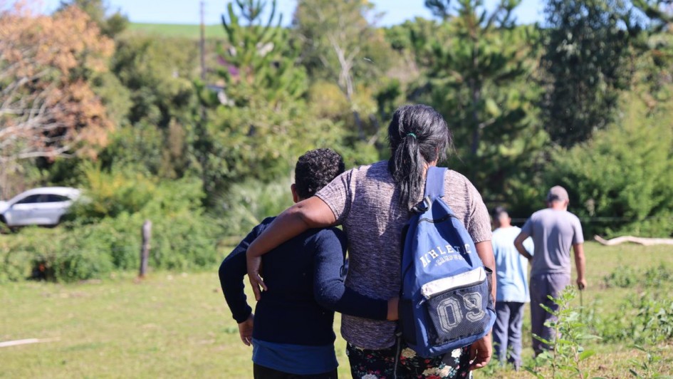 &Aacute;rea externa rural em dia ensolarado, com vegeta&ccedil;&atilde;o abundante e &aacute;rvores ao redor, onde um grupo de pessoas caminha por um terreno gramado. Em primeiro plano, duas pessoas s&atilde;o vistas de costas caminhando lado a lado com os bra&ccedil;os apoiados nos ombros uma da outra, enquanto outras seguem &agrave; frente.