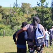 &Aacute;rea externa rural em dia ensolarado, com vegeta&ccedil;&atilde;o abundante e &aacute;rvores ao redor, onde um grupo de pessoas caminha por um terreno gramado. Em primeiro plano, duas pessoas s&atilde;o vistas de costas caminhando lado a lado com os bra&ccedil;os apoiados nos ombros uma da outra, enquanto outras seguem &agrave; frente.