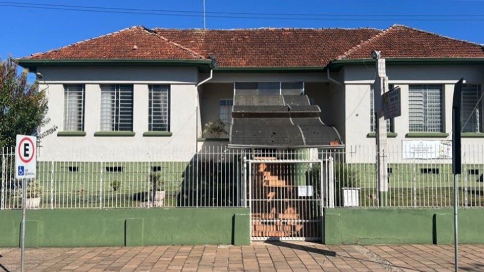 Fachada frontal de um pr&eacute;dio baixo com estilo simples e sim&eacute;trico, paredes claras com detalhes em verde e um telhado inclinado de telhas cer&acirc;micas avermelhadas; o acesso central possui uma escadaria coberta e &eacute; protegido por uma grade met&aacute;lica branca.