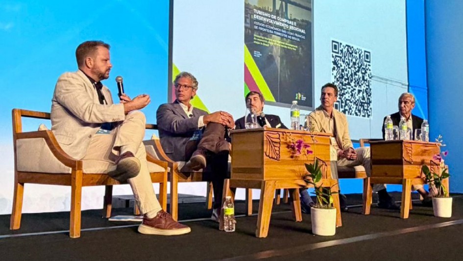 Cinco pessoas est&atilde;o sentadas em cadeiras em um palco, uma delas fala ao microfone, em um ambiente t&iacute;pico de audit&oacute;rio.