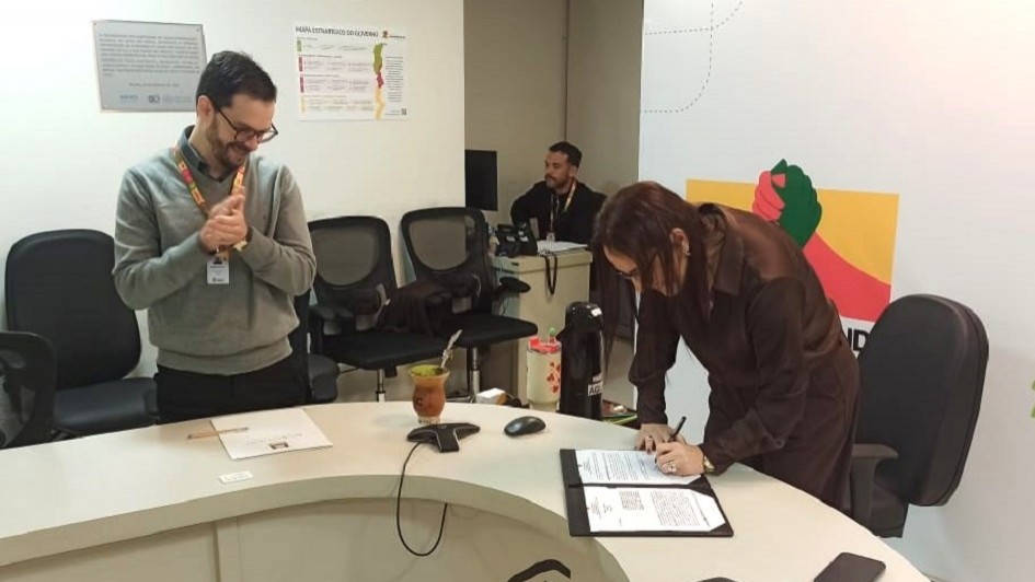 Ambiente de escrit&oacute;rio com balc&atilde;o identificado por &ldquo;RS&rdquo;, onde uma pessoa assina um documento enquanto outra observa ao lado.