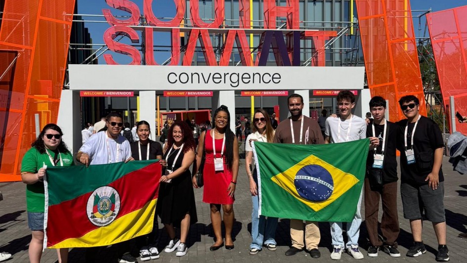 Grupo de pessoas posando em &aacute;rea externa diante de uma estrutura com o letreiro &ldquo;SOUTH SUMMIT&rdquo; e &ldquo;convergence&rdquo;, segurando bandeiras do Brasil e do Rio Grande do Sul; o local tem estruturas modernas com pain&eacute;is coloridos e circula&ccedil;&atilde;o de outras pessoas ao fundo.