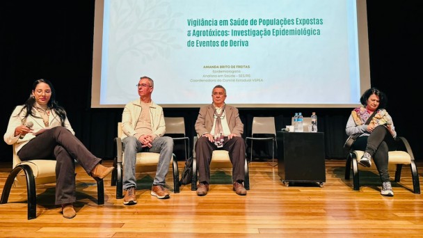 Foto mostra cinco debatedores, sentados em poltronas no palco do evento. No alto, uma tela projeta o nome do evento.
