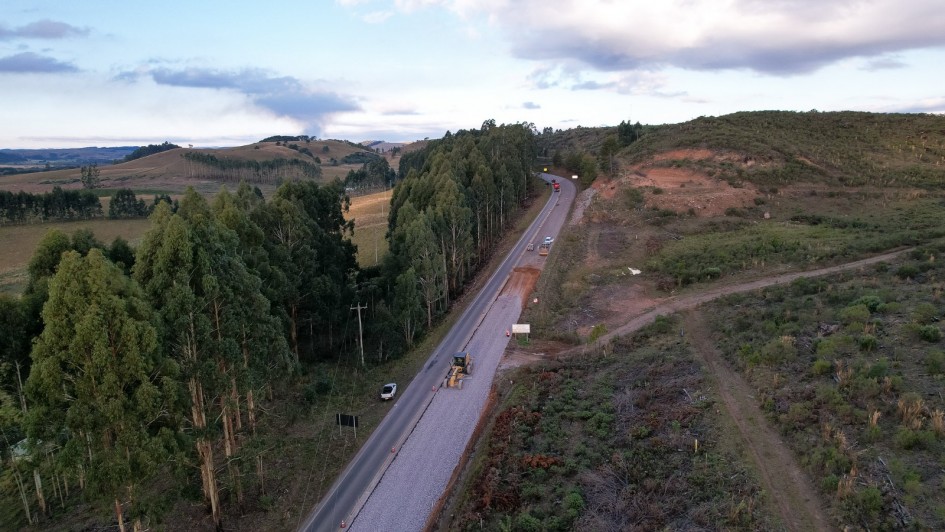Vista a&eacute;rea de uma rodovia em &aacute;rea rural com vegeta&ccedil;&atilde;o e morros ao redor, onde um trecho est&aacute; em obras com m&aacute;quinas e ve&iacute;culos, incluindo caminh&otilde;es e equipamentos ao longo da pista parcialmente preparada.