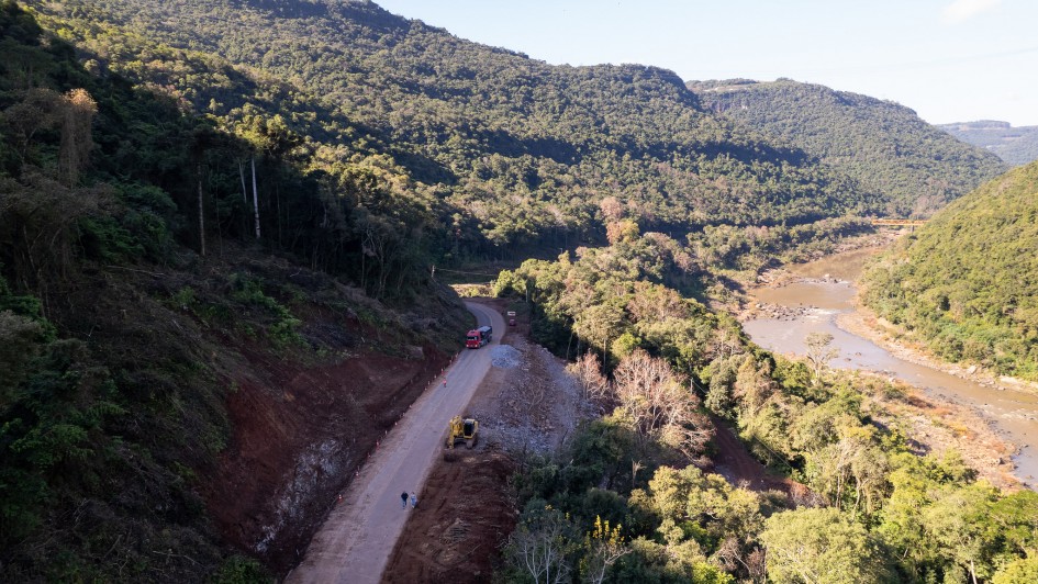 A fotografia em plano panor&acirc;mico e &acirc;ngulo elevado captura uma estrada sinuosa que contorna a encosta de um morro, margeando um rio caudaloso em uma vasta regi&atilde;o montanhosa e vales verdejantes.

A Rodovia e as Obras (Centro-Esquerda):

Uma estrada estreita de asfalto claro corta a imagem diagonalmente a partir do canto inferior esquerdo em dire&ccedil;&atilde;o ao centro.

&Agrave; esquerda da pista, a encosta do morro apresenta trechos de terra roxa e encostas escavadas, evidenciando marcas de deslizamentos anteriores ou trabalhos de terraplanagem. Cones de sinaliza&ccedil;&atilde;o vermelhos e brancos est&atilde;o perfilados na margem para isolar a pista.

No trecho inferior da estrada, uma pequena escavadeira amarela est&aacute; posicionada na margem direita da via, onde h&aacute; um ac&uacute;mulo de terra batida e pedras. Pr&oacute;ximo a ela, duas pessoas caminham a p&eacute;. Mais &agrave; frente, na curva da estrada, um caminh&atilde;o de carga de cor vermelha segue viagem.

O Rio e o Vale (Direita): Correndo paralelamente &agrave; rodovia, um grande rio de &aacute;guas barrentas em tom marrom-claro serpenteia pelo vale &agrave; direita. Suas margens s&atilde;o irregulares e cobertas por pedregais e vegeta&ccedil;&atilde;o rasteira. Ao longe, no canto superior direito, nota-se uma ponte que cruza o leito do rio.

A Paisagem e o Relevo:

A paisagem &eacute; imponente e dominada por grandes montanhas e morros recobertos por uma floresta densa e cont&iacute;nua de Mata Atl&acirc;ntica em tons de verde-escuro.

A ilumina&ccedil;&atilde;o solar vem da direita, projetando sombras longas das &aacute;rvores e do pr&oacute;prio relevo sobre a encosta e a estrada no lado esquerdo, sugerindo o in&iacute;cio da manh&atilde; ou o final da tarde. O c&eacute;u na parte superior est&aacute; limpo e claro.

A imagem registra a complexidade geogr&aacute;fica e os desafios de infraestrutura e engenharia para a manuten&ccedil;&atilde;o de rodovias em regi&otilde;es de serra.
