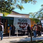 &Aacute;rea externa urbana com uma unidade m&oacute;vel identificada pelo texto &ldquo;TUDO F&Aacute;CIL&rdquo; estacionada junto &agrave; cal&ccedil;ada, onde v&aacute;rias pessoas est&atilde;o reunidas formando uma pequena fila pr&oacute;xima &agrave; entrada.