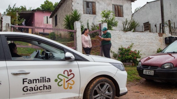 Foto mostra ve&iacute;culo com identifica&ccedil;&atilde;o do Programa Fam&iacute;lia Ga&uacute;cha parado na frente de uma casa. Uma agente, vestida de camiseta e bon&eacute; verde do programa anota na prancheta enquanto conversa com uma mulher no port&atilde;o da casa. 