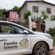 Foto mostra ve&iacute;culo com identifica&ccedil;&atilde;o do Programa Fam&iacute;lia Ga&uacute;cha parado na frente de uma casa. Uma agente, vestida de camiseta e bon&eacute; verde do programa anota na prancheta enquanto conversa com uma mulher no port&atilde;o da casa. 