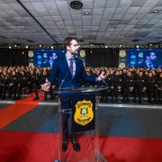 O governador discursa em p&uacute;lpito transparente com o bras&atilde;o &ldquo;Pol&iacute;cia Penal RS&rdquo;, diante de uma plateia extensa de participantes uniformizados, com tel&otilde;es replicando a cena e ilumina&ccedil;&atilde;o c&ecirc;nica no ambiente.