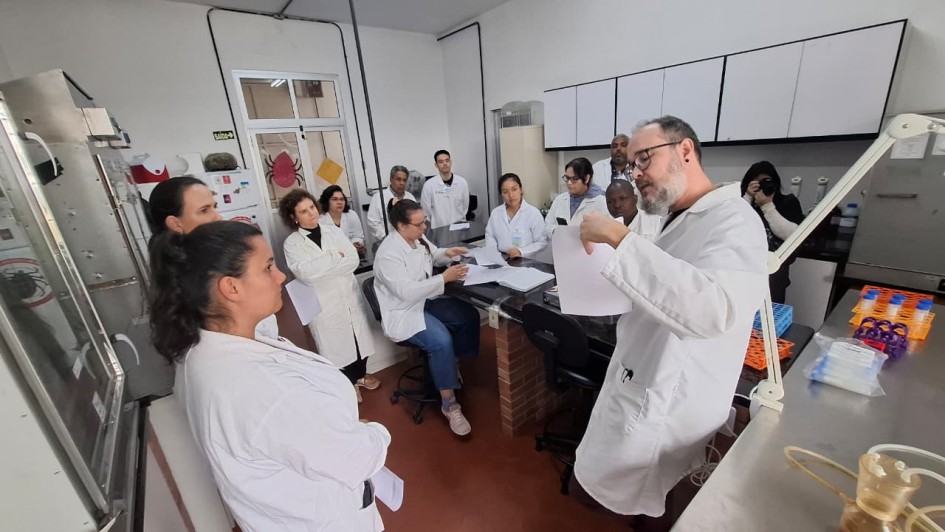 Fotografia mostra um grupo de cerca de 12 pessoas usando jalecos brancos reunidas em um laborat&oacute;rio. Em primeiro plano, um homem de jaleco gesticula enquanto segura folhas de papel e fala aos participantes, que o observam atentamente. Ao redor, alguns integrantes est&atilde;o em p&eacute; e outros sentados junto a uma bancada central. O ambiente cont&eacute;m equipamentos laboratoriais, arm&aacute;rios suspensos, pipetas, suportes para tubos de ensaio e outros materiais de pesquisa, indicando uma atividade de treinamento ou orienta&ccedil;&atilde;o t&eacute;cnica.