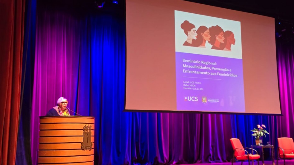 A fotografia registra a realiza&ccedil;&atilde;o de um evento institucional em um audit&oacute;rio, identificado pelo nome do arquivo e pelo slide projetado como o "Semin&aacute;rio Regional: Masculinidades, Preven&ccedil;&atilde;o e Enfrentamento aos Feminic&iacute;dios".

O Palco e a Palestrante
A&ccedil;&atilde;o Principal: No lado esquerdo, uma mulher de cabelos curtos e claros, usando &oacute;culos de arma&ccedil;&atilde;o vermelha e um len&ccedil;o colorido, fala ao microfone posicionada atr&aacute;s de um p&uacute;lpito cil&iacute;ndrico de madeira com detalhes em frisos horizontais.

Mobili&aacute;rio do Palco: No lado direito do palco, sobre um tapete vermelho, h&aacute; duas poltronas estofadas tamb&eacute;m na cor vermelha e uma pequena mesa de centro de vidro com um arranjo de flores (ant&uacute;rios), configurando um espa&ccedil;o para debates.

Elementos de Proje&ccedil;&atilde;o
Tel&atilde;o Central: Uma grande tela de proje&ccedil;&atilde;o est&aacute; centralizada no palco, exibindo a identidade visual do evento. Na parte superior do slide, h&aacute; uma ilustra&ccedil;&atilde;o estilizada com o perfil de quatro mulheres de diferentes etnias.

Texto do Slide: Logo abaixo da ilustra&ccedil;&atilde;o, o slide em tom lil&aacute;s traz o t&iacute;tulo do semin&aacute;rio e informa&ccedil;&otilde;es institucionais, incluindo o local (UCS Teatro), a data (10/06) e o hor&aacute;rio (09h &agrave;s 18h). No rodap&eacute; do slide, destacam-se os logotipos da UCS (Universidade de Caxias do Sul) e do Governo do Estado do Rio Grande do Sul.

Ambiente e Ilumina&ccedil;&atilde;o
Cen&aacute;rio: O fundo do palco &eacute; composto por grandes cortinas de tecido que recebem uma ilumina&ccedil;&atilde;o c&ecirc;nica marcante em tons de azul e violeta. A ilumina&ccedil;&atilde;o cria um forte contraste com o piso de madeira do palco e a estrutura c&ecirc;nica superior.

Perspectiva: A foto foi tirada a partir da plateia, ligeiramente de baixo para cima, mostrando o topo da cabe&ccedil;a de um espectador em primeiro plano no canto inferior direito.