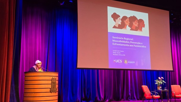 A fotografia registra a realiza&ccedil;&atilde;o de um evento institucional em um audit&oacute;rio, identificado pelo nome do arquivo e pelo slide projetado como o "Semin&aacute;rio Regional: Masculinidades, Preven&ccedil;&atilde;o e Enfrentamento aos Feminic&iacute;dios".

O Palco e a Palestrante
A&ccedil;&atilde;o Principal: No lado esquerdo, uma mulher de cabelos curtos e claros, usando &oacute;culos de arma&ccedil;&atilde;o vermelha e um len&ccedil;o colorido, fala ao microfone posicionada atr&aacute;s de um p&uacute;lpito cil&iacute;ndrico de madeira com detalhes em frisos horizontais.

Mobili&aacute;rio do Palco: No lado direito do palco, sobre um tapete vermelho, h&aacute; duas poltronas estofadas tamb&eacute;m na cor vermelha e uma pequena mesa de centro de vidro com um arranjo de flores (ant&uacute;rios), configurando um espa&ccedil;o para debates.

Elementos de Proje&ccedil;&atilde;o
Tel&atilde;o Central: Uma grande tela de proje&ccedil;&atilde;o est&aacute; centralizada no palco, exibindo a identidade visual do evento. Na parte superior do slide, h&aacute; uma ilustra&ccedil;&atilde;o estilizada com o perfil de quatro mulheres de diferentes etnias.

Texto do Slide: Logo abaixo da ilustra&ccedil;&atilde;o, o slide em tom lil&aacute;s traz o t&iacute;tulo do semin&aacute;rio e informa&ccedil;&otilde;es institucionais, incluindo o local (UCS Teatro), a data (10/06) e o hor&aacute;rio (09h &agrave;s 18h). No rodap&eacute; do slide, destacam-se os logotipos da UCS (Universidade de Caxias do Sul) e do Governo do Estado do Rio Grande do Sul.

Ambiente e Ilumina&ccedil;&atilde;o
Cen&aacute;rio: O fundo do palco &eacute; composto por grandes cortinas de tecido que recebem uma ilumina&ccedil;&atilde;o c&ecirc;nica marcante em tons de azul e violeta. A ilumina&ccedil;&atilde;o cria um forte contraste com o piso de madeira do palco e a estrutura c&ecirc;nica superior.

Perspectiva: A foto foi tirada a partir da plateia, ligeiramente de baixo para cima, mostrando o topo da cabe&ccedil;a de um espectador em primeiro plano no canto inferior direito.