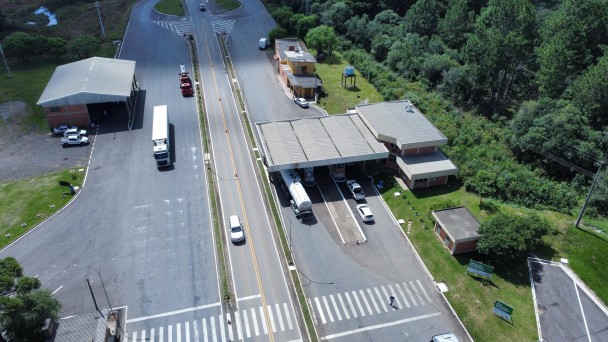 Esta &eacute; uma descri&ccedil;&atilde;o da imagem para fins de acessibilidade:

Uma fotografia a&eacute;rea, capturada por um drone em &acirc;ngulo picado (de cima para baixo), mostra a estrutura do Posto Fiscal de Vacaria, localizado &agrave;s margens de uma rodovia duplicada.

Estrutura Principal e Rodovia
O Posto Fiscal: No centro da imagem, destaca-se uma grande cobertura met&aacute;lica arqueada e alongada, pintada de branco com detalhes em azul nas bordas frontais. Sob essa estrutura, encontram-se as pistas de triagem e pesagem para os ve&iacute;culos de carga.

A Rodovia: A estrada possui pistas duplicadas em asfalto escuro, separadas por uma barreira central de concreto (tipo New Jersey) e faixas sinalizadoras pintadas em amarelo e branco. O fluxo de ve&iacute;culos &eacute; moderado, observando-se alguns caminh&otilde;es e carros transitando ou estacionados nas proximidades do posto.

Edifica&ccedil;&otilde;es de Apoio: &Agrave; esquerda da grande cobertura, h&aacute; pr&eacute;dios administrativos de apoio com telhados em tons de cinza e &aacute;reas de estacionamento interno para ve&iacute;culos oficiais e de funcion&aacute;rios.

Ambiente e Entorno
Vegeta&ccedil;&atilde;o: A infraestrutura rodovi&aacute;ria &eacute; cercada por amplas &aacute;reas verdes. H&aacute; gramados bem cuidados nas margens e canteiros, al&eacute;m de copas de &aacute;rvores nativas dispersas ao redor do complexo.

Ilumina&ccedil;&atilde;o e Clima: A imagem foi registrada em um dia claro, com luz solar direta que projeta sombras n&iacute;tidas das &aacute;rvores, dos ve&iacute;culos e da pr&oacute;pria estrutura met&aacute;lica sobre o solo. Ao fundo, a paisagem se estende em &aacute;reas de campo aberto sob uma linha de horizonte suave.